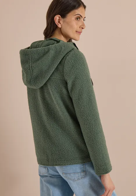 Frau trägt grünen Teddy-Fleece-Hoodie mit Kapuze und Jeans, Rückenansicht vor beigem Hintergrund.