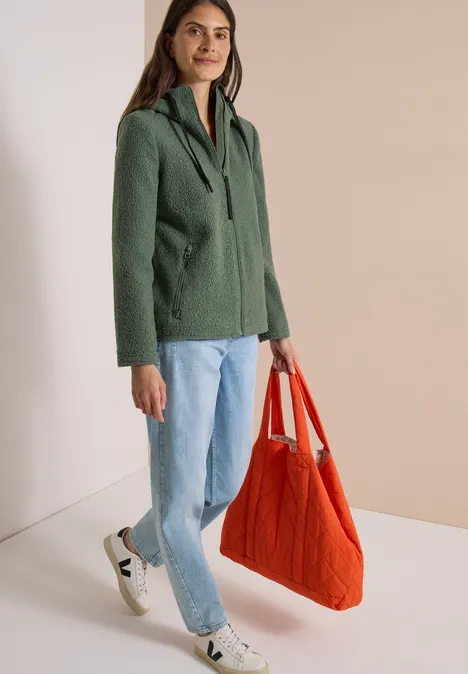 Frau trägt grünen Kapuzenblazer, helle Jeans, weiße Sneaker und hält rote Stofftasche vor neutralem Hintergrund.