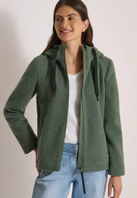 Frau trägt grüne Teddy-Fleecejacke mit Kapuze, weiße Bluse, lächelt seitlich, Hände in den Taschen.