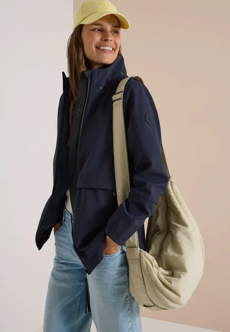 Junge Frau lächelt, trägt eine Cap, eine dunkelblaue Jacke, Jeans und eine große Tasche über der Schulter.