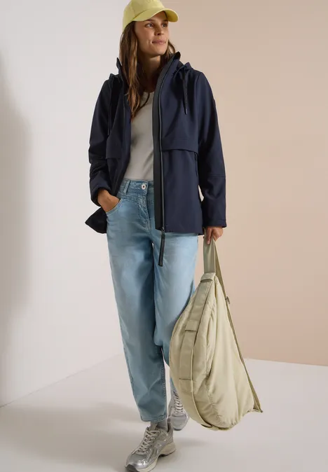 Eine Frau trägt eine dunkle Jacke, weiße Bluse, weite Jeans, weiße Sneaker, gelbe Kappe und eine gesteppte Tasche.
