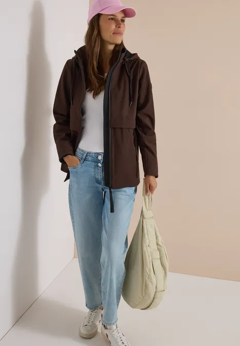 Junge Frau in brauner Jacke, weißem T-Shirt, hellblauen Jeans, rosa Cap und Canvas-Beutel.