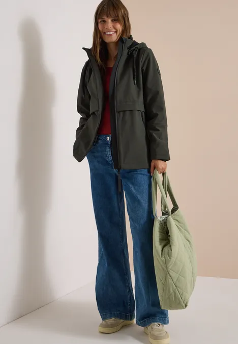 Frau in grüner Jacke, rotem Pullover, Jeans und Sneakern, hält grüne Stepp-Tasche vor neutralem Hintergrund.