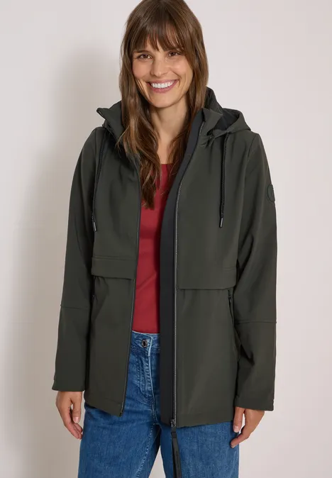 Eine Frau lächelt und trägt eine grüne Jacke über einem roten Shirt und Jeans vor neutralem Hintergrund