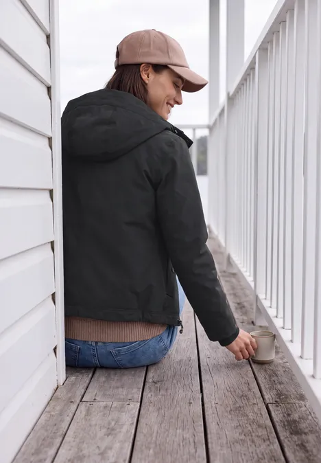 Kurze Jacke mit 2-in-1 Kapuze Black Frau in schwarzer Jacke, blauer Jeans und Cap sitzt auf Holzterrasse, lehnt ans Geländer und lächelt.