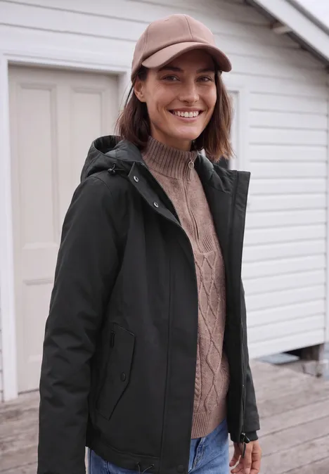 Kurze Jacke mit 2-in-1 Kapuze Black Frau mit Baseballcap, Strickpullover und schwarzer Jacke lächelt vor einem hellen Gebäude.
