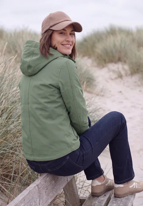 Kurze Jacke mit 2-in-1 Kapuze shadow khaki Frau in grüner Jacke und Jeans sitzt auf Holzbalken am sandigen Dünenstrand und schaut lächelnd zurück.