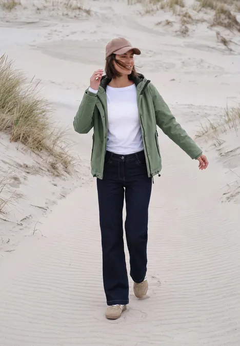 Kurze Jacke mit 2-in-1 Kapuze shadow khaki Eine Frau in grüner Jacke, Jeans und Cap spaziert über Dünen.