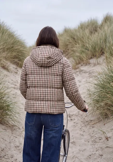 Eine Person in kariertem Parka schiebt ein Fahrrad zwischen grasigen Dünen am Strand.