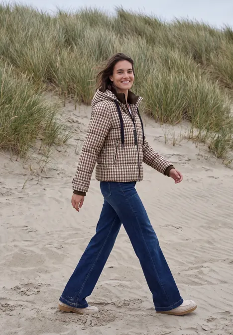 Eine Frau in kariertem Mantel und Schlagjeans spaziert entspannt über grasbewachsene Dünen.