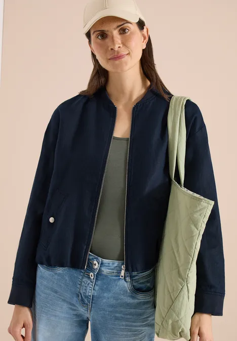 Blouson aus Leinen mit Zipper universal blue Frau in dunkler Jacke, grünem Top, Jeans und hellgrüner Stepp-Tasche über der Schulter.