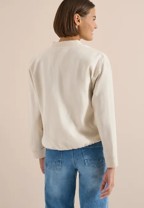 Person von hinten mit hellem Pullover und blauer Jeans vor neutralem Hintergrund.