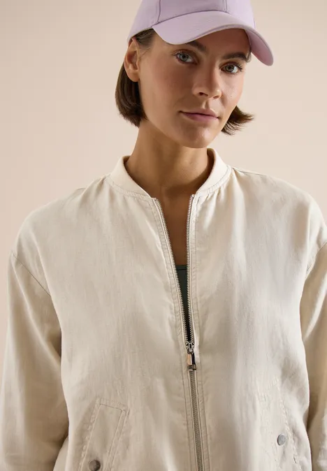 Frau in hellbeiger Jacke mit Reißverschluss und rosa Baseballcap vor neutralem Hintergrund.