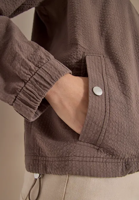 Braune, strukturierte Jacke mit aufgesetzter Tasche und Metallknopf, Hand steckt in der Tasche.