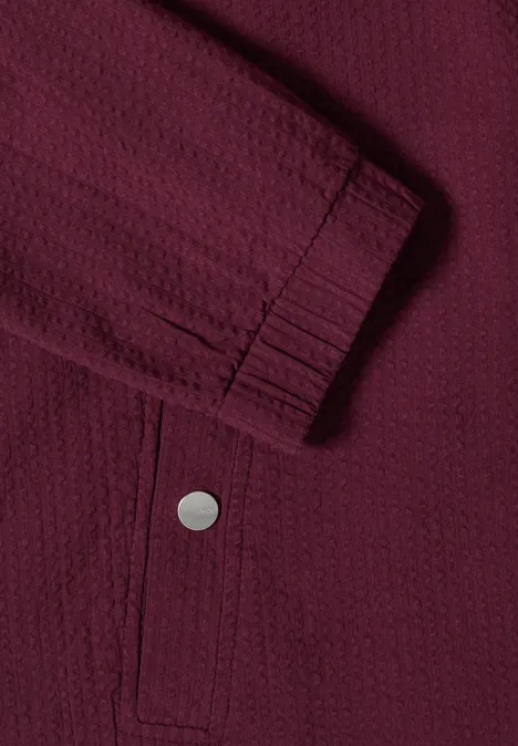 Nahaufnahme einer dunkelroten Jacke mit strukturiertem Stoff, elastischem Ärmelbündchen und Druckknopf-Tasche.