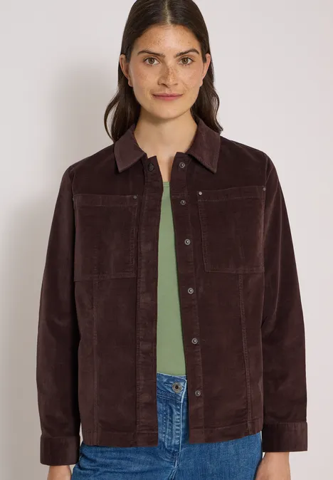 Femme en veste velours marron, top vert et jean, souriant légèrement sur fond clair.