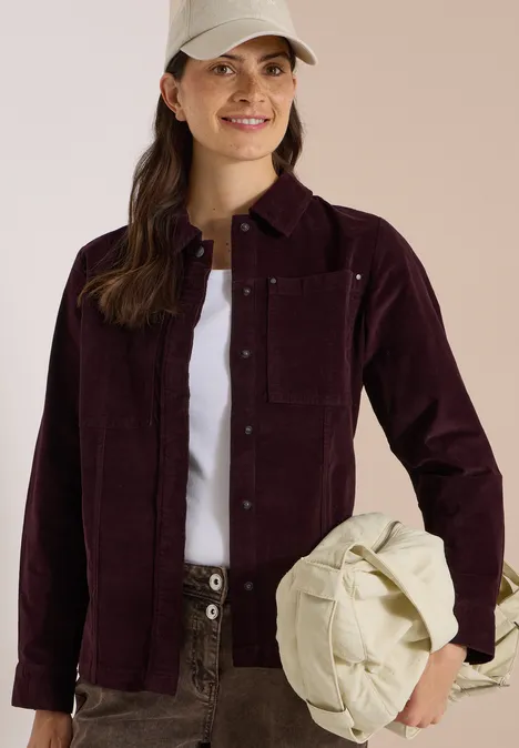 Femme souriante en veste en velours côtelé bordeaux, t-shirt blanc et pantalon vert, tenant un sac crème.