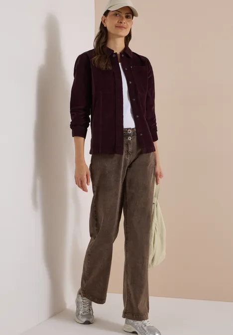 Femme en look décontracté : top blanc, veste bordeaux, pantalon velours marron, baskets argentées, casquette.