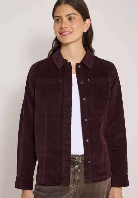 Une jeune femme porte une veste en velours côtelé violet foncé sur un t-shirt blanc et un pantalon à motifs.