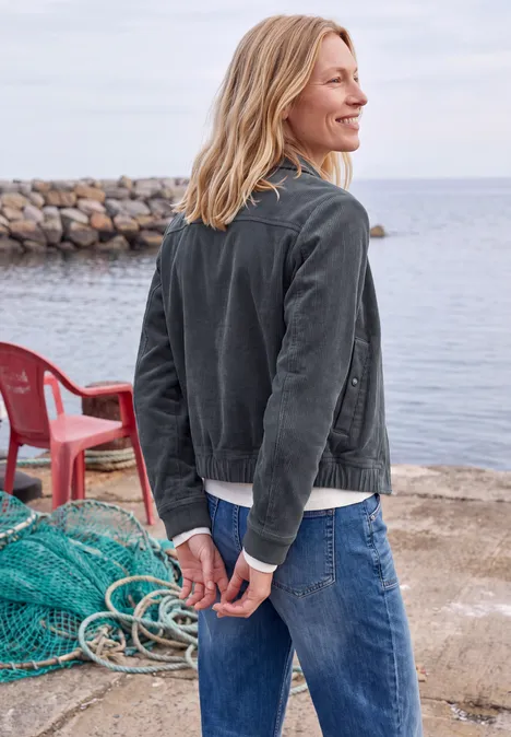 Femme en veste en jean, dos tourné, mains dans le dos, face à la mer, rochers et chaises rouges en arrière-plan.