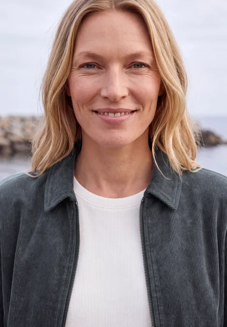 Femme blonde souriante sur la plage, portant un t-shirt blanc et une veste grise.