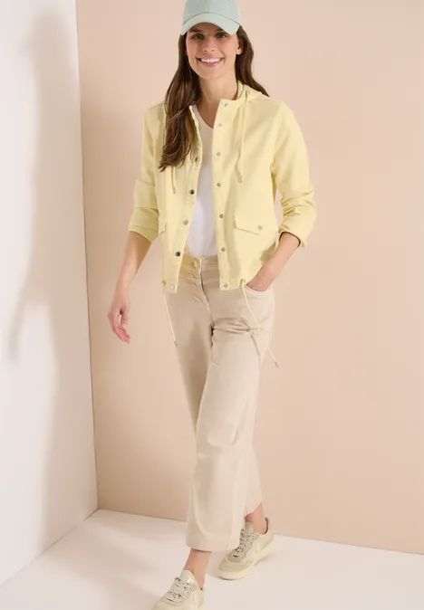 Frau steht in hellem Raum, trägt gelbe Jacke, weißes Top, beige Hose und Sneaker.