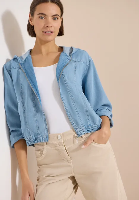 Persoon met lichtblauw spijkerjack en beige broek tegen neutrale achtergrond.