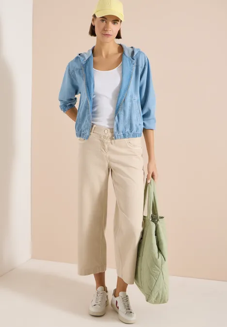 Een vrouw draagt een wit t-shirt, een lichtblauw jasje, beige wijde broek, witte sneakers en een groene tas.
