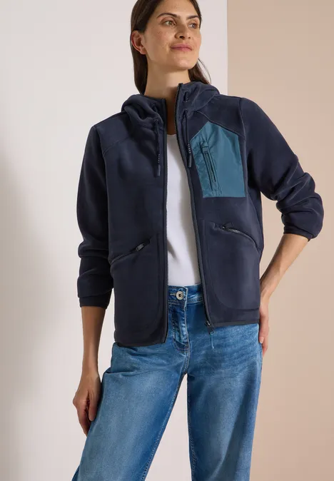 Eine Frau trägt eine dunkelblaue Fleecejacke mit Kapuze, Reißverschluss und kontrastfarbener Brusttasche.