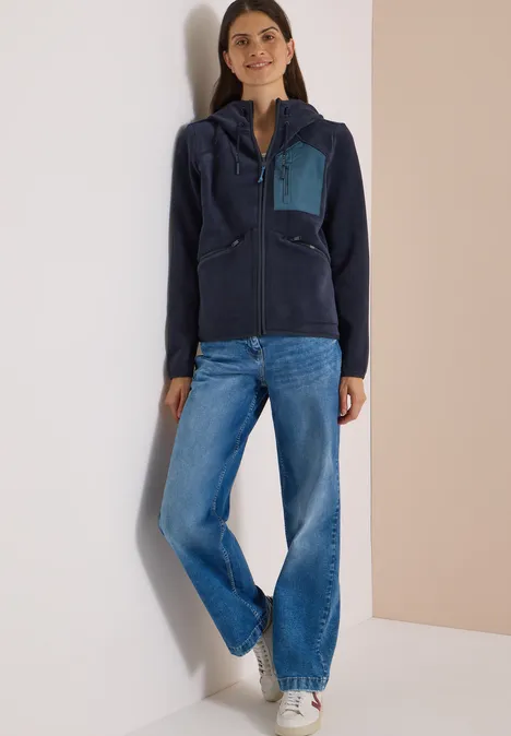 Frau lehnt an Wand, trägt dunkelblaue Fleecejacke mit hellblauer Brusttasche, weite Jeans und weiße Sneaker.
