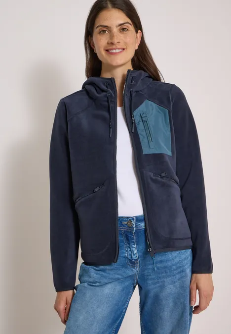 Frau trägt dunkelblaue Fleecejacke mit Kapuze, weißes Top und Jeans vor hellem Hintergrund.