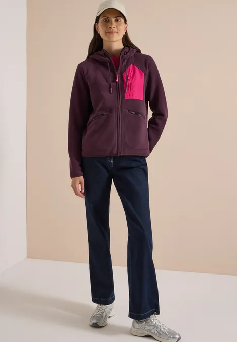 Frau in lila Jacke mit pinker Tasche, blauen Jeans, grauen Sneakern und Cap vor hellem Hintergrund.