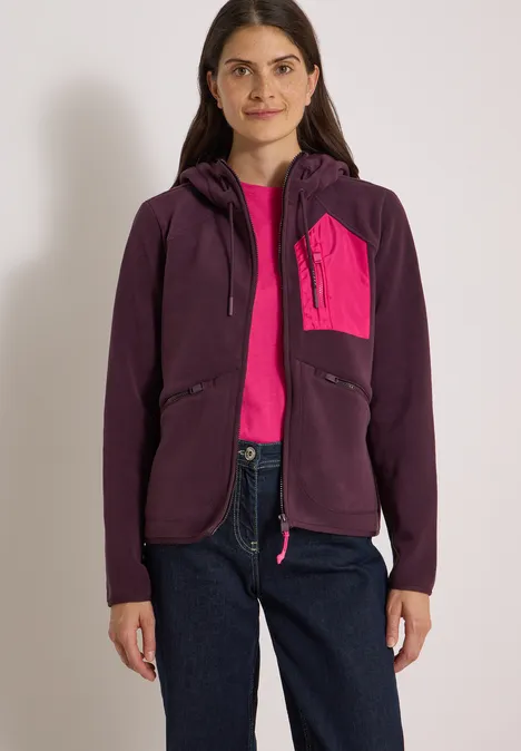 Frau in lila Kapuzenjacke mit pinker Brusttasche, pinkem Shirt und Jeans vor neutralem Hintergrund.