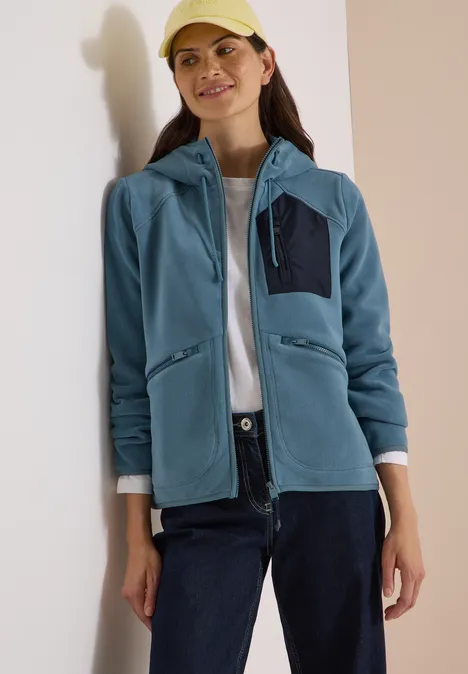Vrouw in blauw fleece-hoodie, wit shirt, donkere broek en pet voor een neutrale achtergrond.