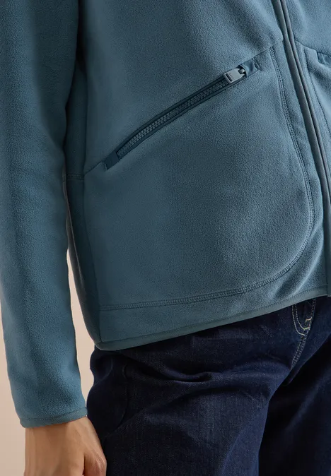 Persoon draagt turquoise fleecevest met ritssluiting en blauwe spijkerbroek.