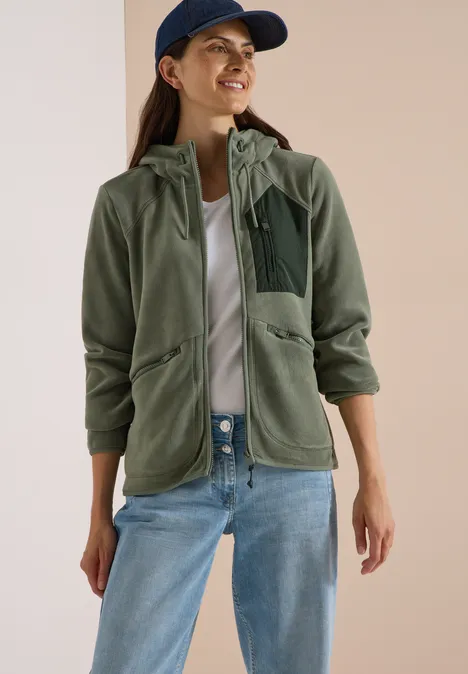 Vrouw met groene fleecevest met capuchon, witte top, spijkerbroek en pet.