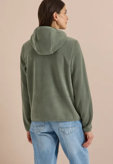 Vrouw van achteren in groen met capuchon en spijkerbroek, kijkt iets opzij
