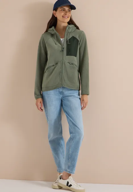 Vrouw in groene jas, spijkerbroek, sneakers en pet voor een beige achtergrond.