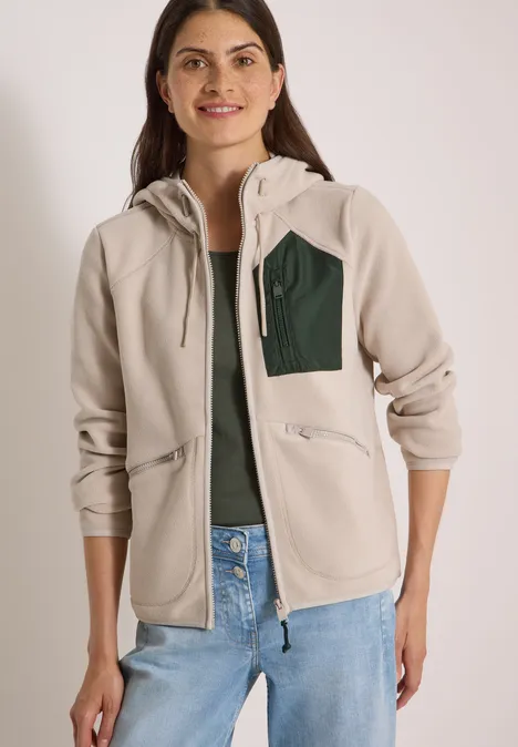 Frau trägt helle Fleece-Jacke mit Kapuze, grünem Brustfach und Jeans