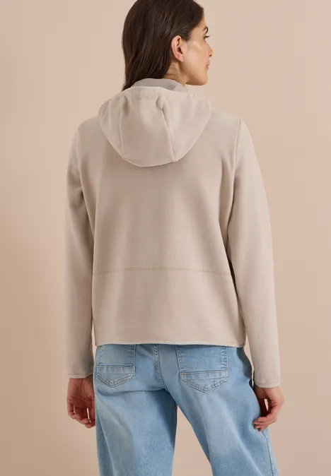 Frau von hinten in hellgrauer Fleece-Jacke mit Kapuze und blauen Jeans vor neutralem Hintergrund.
