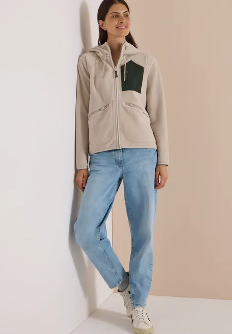 Frau lehnt an Wand in Freizeit-Outfit mit beiger Fleecejacke, Jeans und weißen Sneakern.