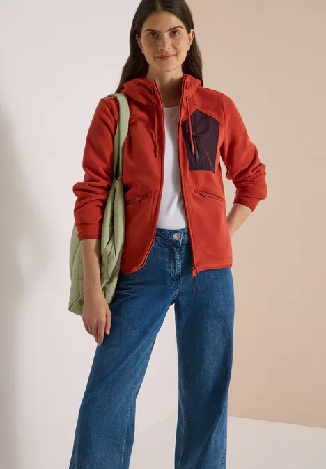 Eine Frau trägt eine rote Jacke, weiße Bluse, blaue Jeans und hält eine grüne Stofftasche.