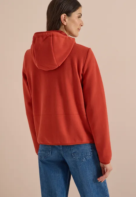 Frau von hinten in roter Kapuzenjacke und blauen Jeans vor einfarbigem Hintergrund.