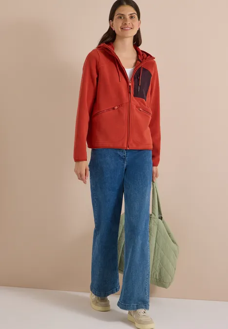 Frau in roter Kapuzenjacke, blauen Jeans und weißen Sneakern hält eine große, grüne Stepp-Tasche vor beigem Hintergrund.