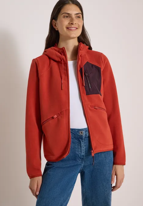 Frau in roter Kapuzenjacke, weißem Shirt und blauen Jeans steht lächelnd.