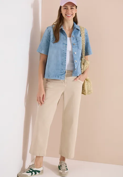 Frau in legerem Outfit: hellblaue Jeansbluse über weißem Top, beigefarbene Culotte, Basttasche und Sneakers.