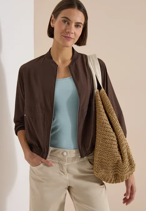 Blouson uni à fermeture zip brown cacao Femme en tenue décontractée: débardeur bleu ciel, veste marron légère, pantalon beige et grand sac tressé.