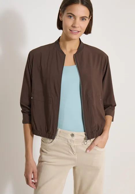 Blouson uni à fermeture zip brown cacao Femme en veste marron légère, top bleu clair et pantalon beige, mains dans les poches.