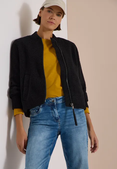 Vrouw tegen muur, zwarte jas, gele trui, blauwe jeans en witte pet.
