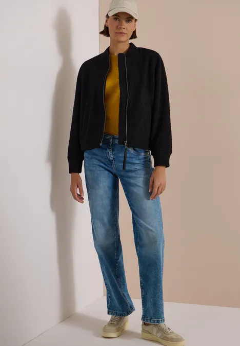 Persoon in een hoek van de kamer, met jeans, geel shirt, zwarte jas, sneakers en pet.